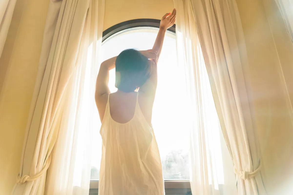 Mujer estirándose frente a una ventana iluminada por la luz cálida de la mañana